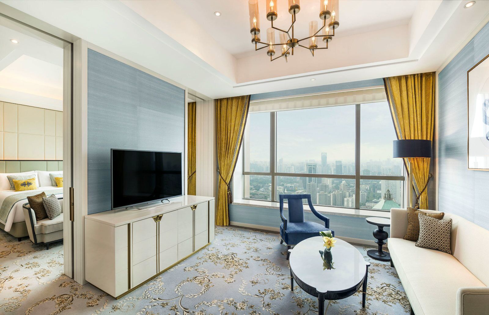 A modern hotel room with a large window offering a city view, a flat-screen TV, a sofa, a dining table, and a blue armchair.