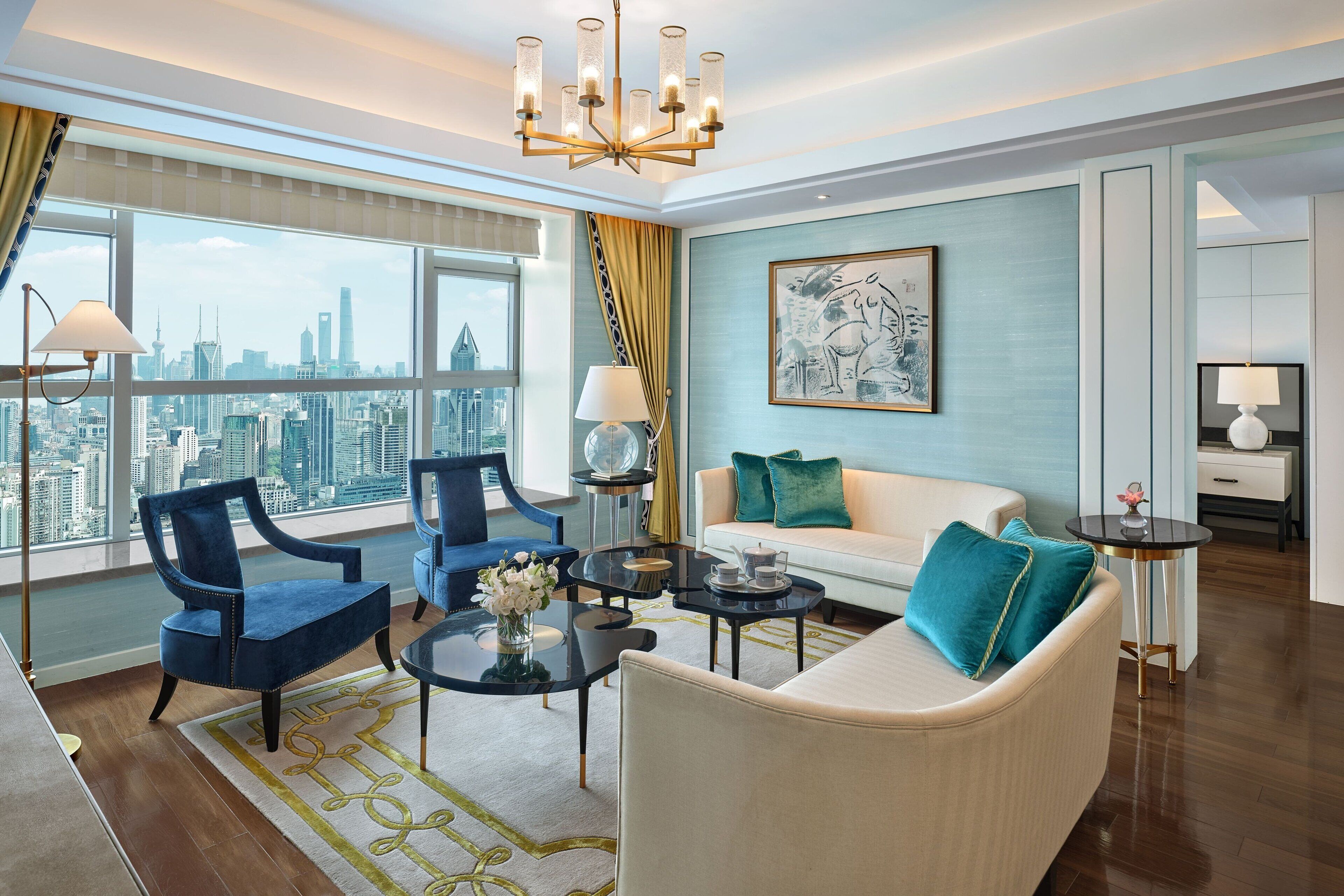 A modern living room with a city view, a chandelier, and a sofa with teal pillows.