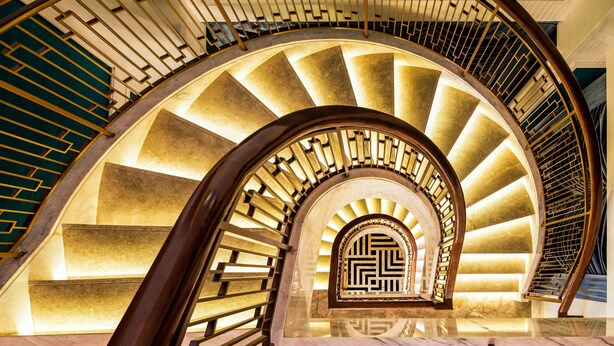Un magnifique escalier en colimaçon, illuminé et orné d’un motif géométrique au sommet, menant à une porte en arc de cercle.