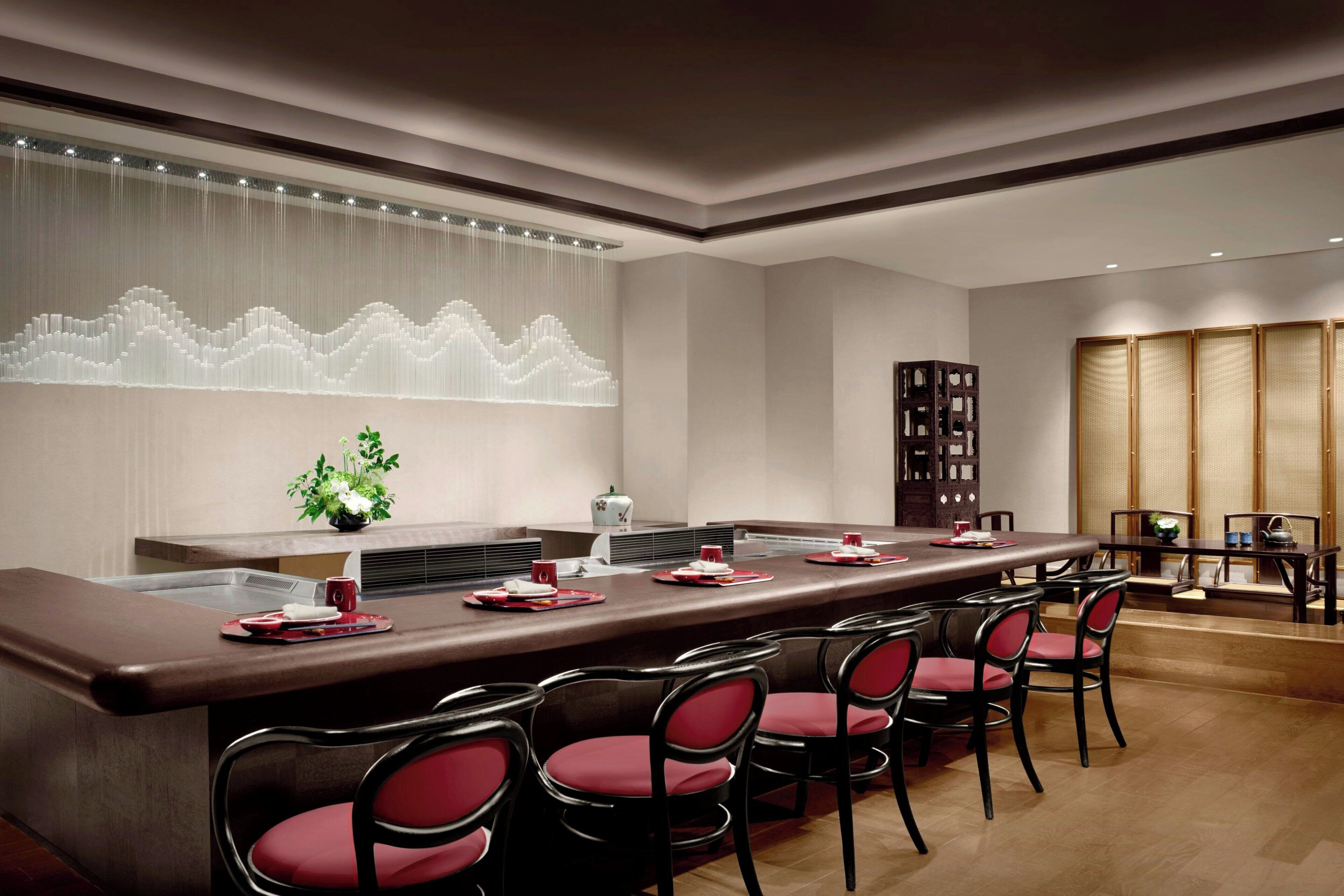 A modern dining area with a long table, red chairs, and a decorative wall feature.