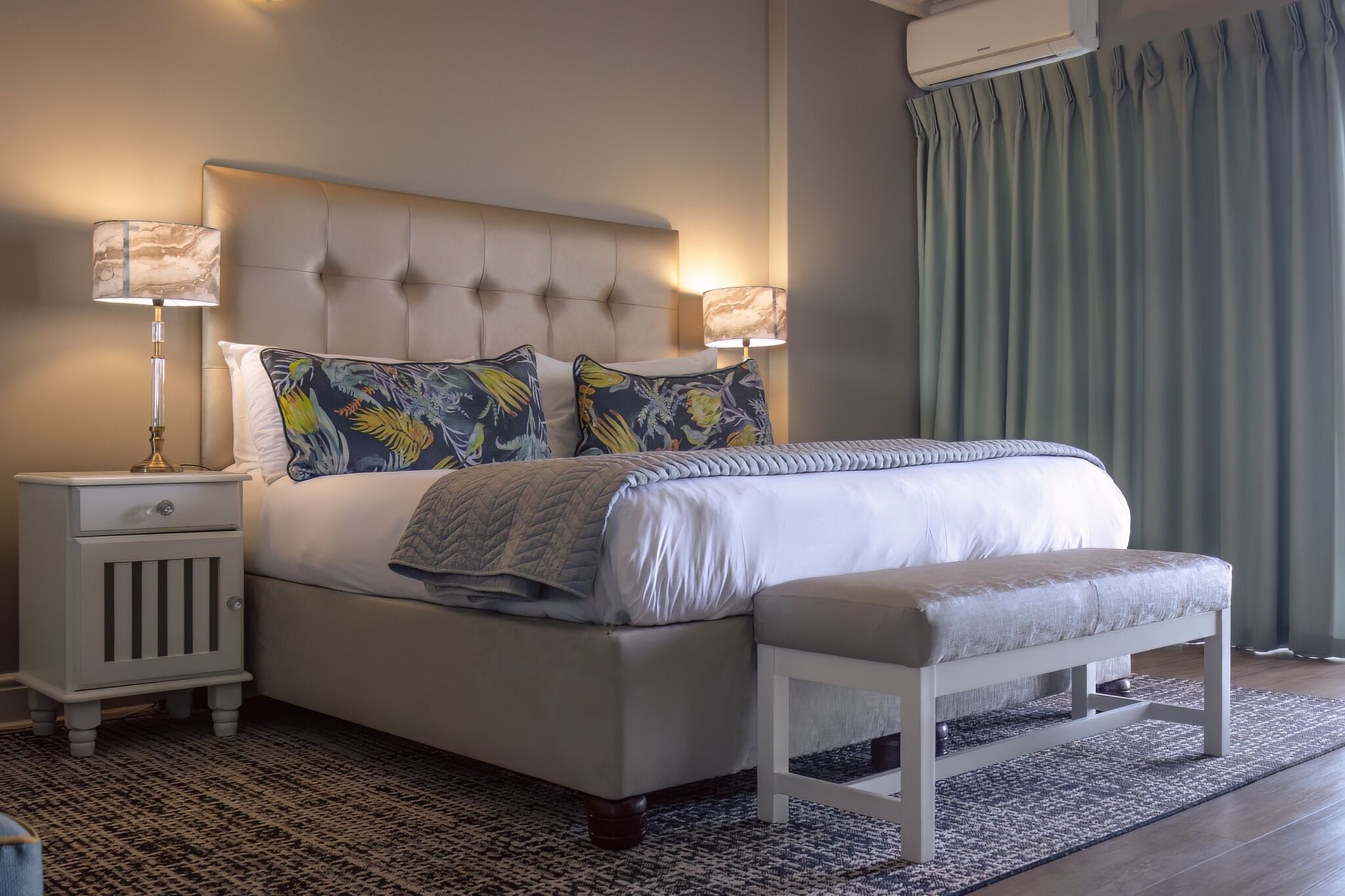 A neatly made bed with a tufted headboard, two decorative pillows, a bedside table with a lamp, and a bench.