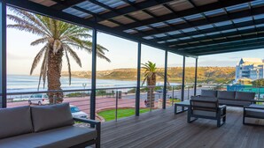 A covered outdoor area with a wooden deck, seating, and a view of the ocean and hills. - Diaz Hotel & Resort (Mossel Bay)