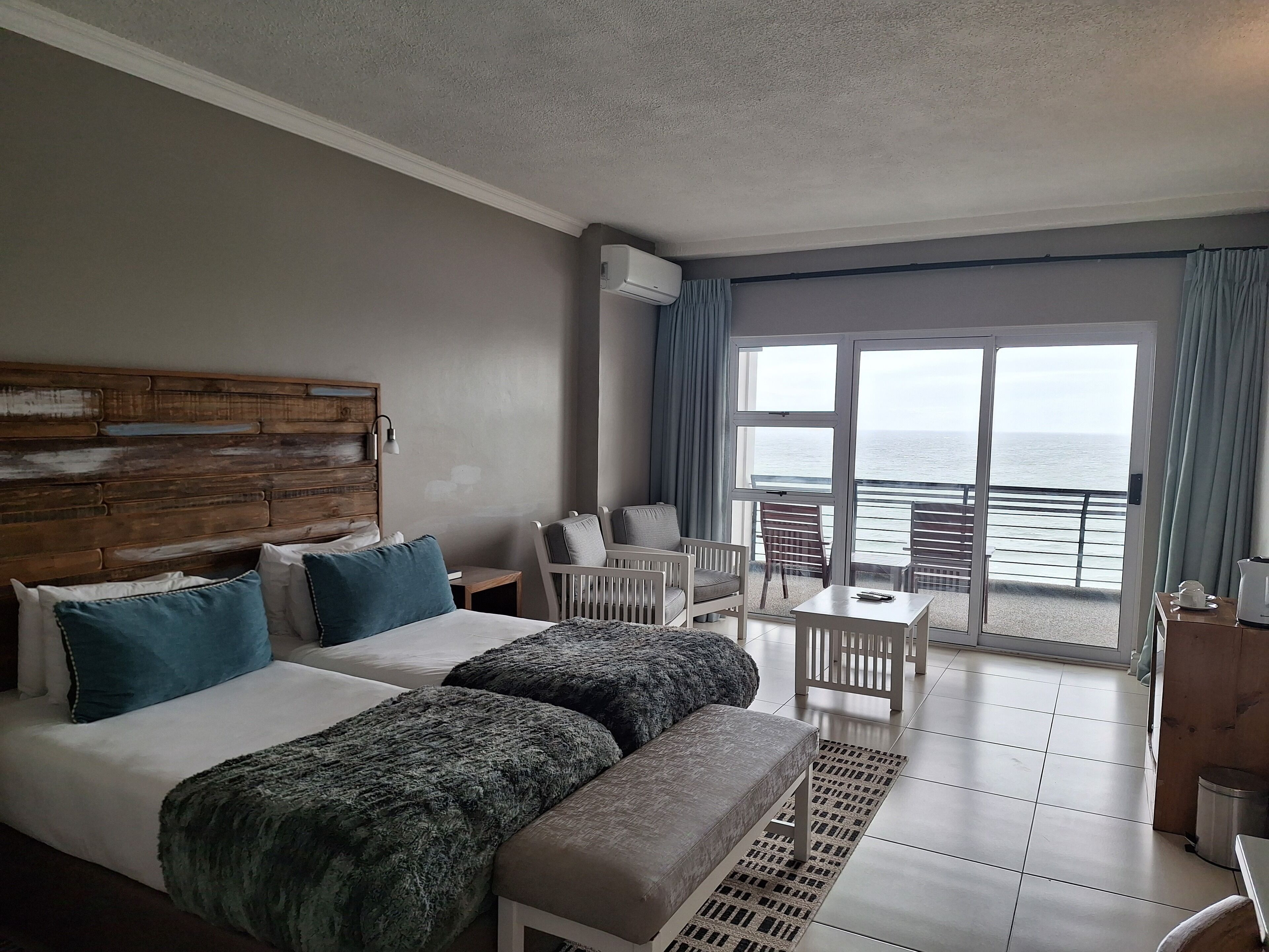 A hotel room with a large bed, two bedside tables, a small table, a chair, and a view of the sea through a sliding door.