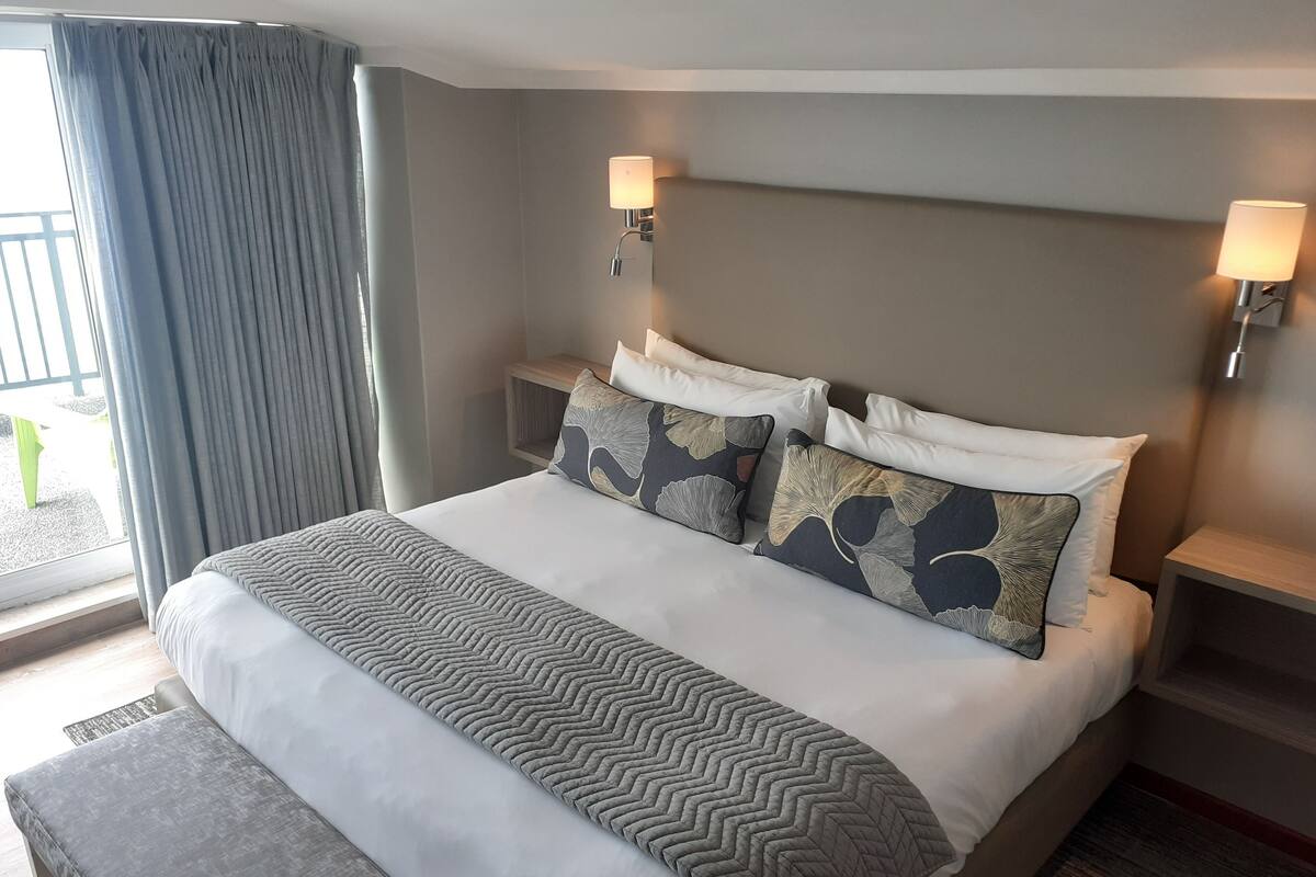 Une chambre d’hôtel avec un grand lit, une table de chevet, une commode, un banc et une vue sur l’extérieur.