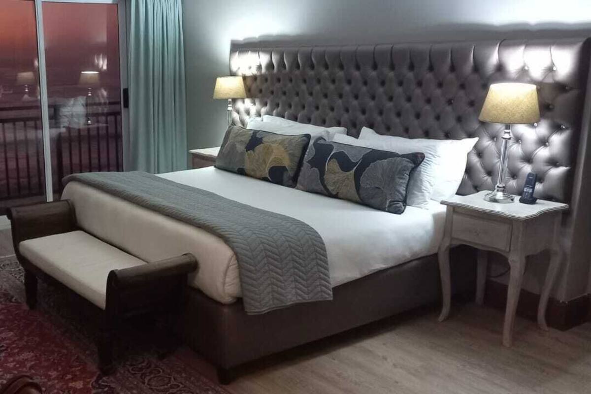Une chambre d’hôtel avec un grand lit, une table de chevet avec une lampe, un banc et une vue sur l’océan par une porte coulissante.