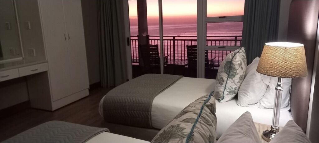 A hotel room with a bed, a nightstand, a lamp, and a view of the ocean at sunset.