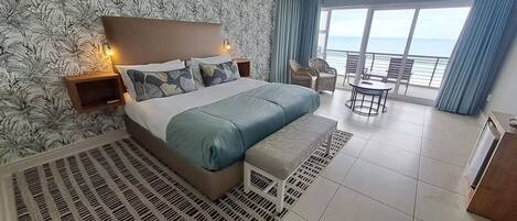A bedroom with a large bed, a nightstand, a chair, and a view of the sea through large windows.