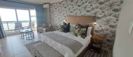 A hotel room with a large bed, a small table, a chair, and a view of the sea through large windows.