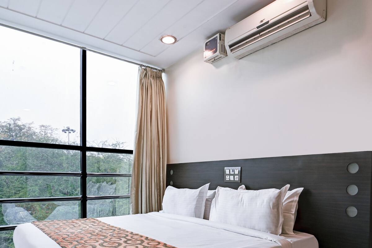 Habitación de hotel con una cama grande, un equipo de aire acondicionado, teléfono junto a la cama y vistas a zonas verdes a través de amplias ventanas.