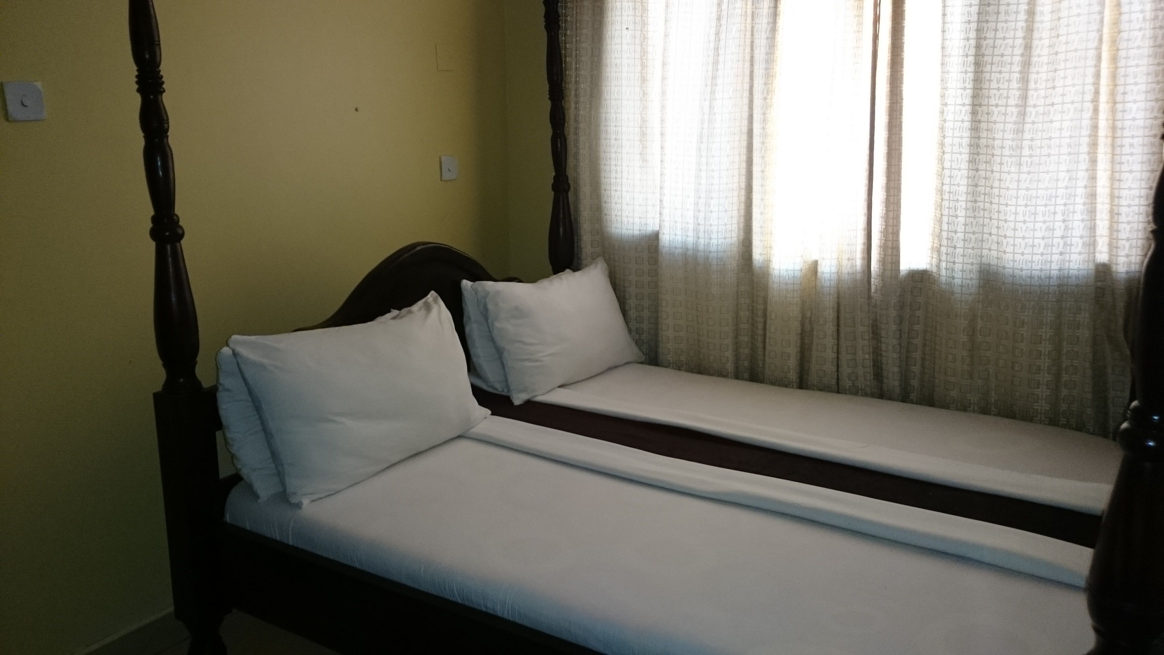 A room with two single beds, a wooden headboard, a window with sheer curtains, and a plain yellow wall.