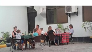An outdoor dining area with tables and chairs, a building with large windows and an air conditioning unit, and potted plants.
