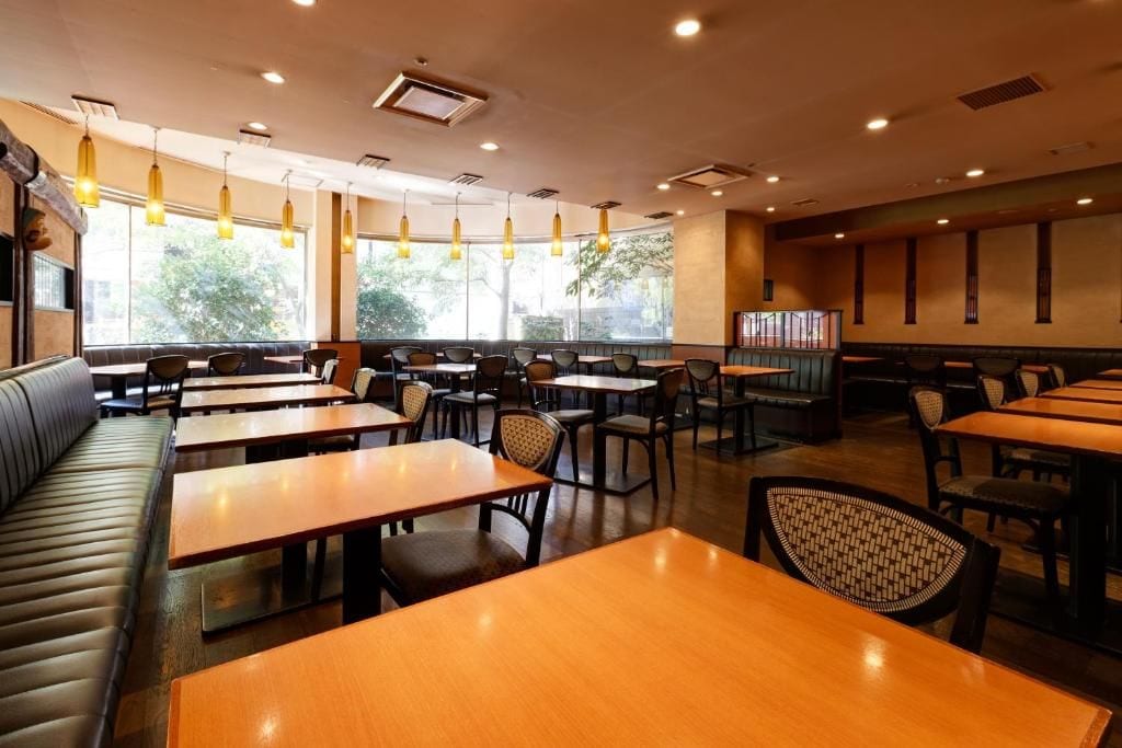 A restaurant interior with multiple tables and booths, pendant lighting, and large windows.