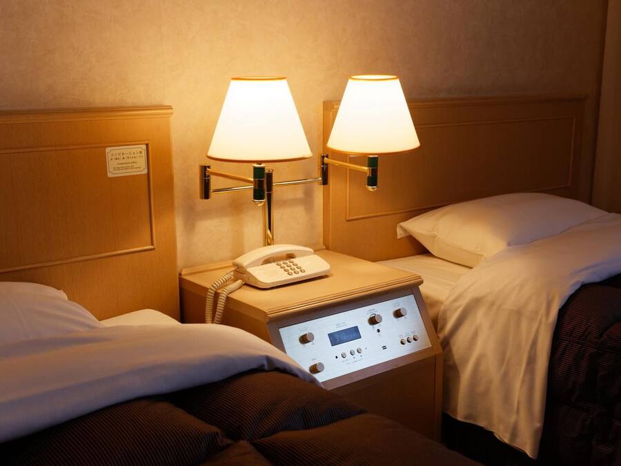 A hotel room with two beds, each with a bedside table and lamp. There is a control panel on the wall between the beds.