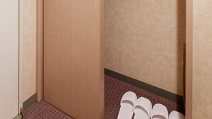Een hotelkamer met een houten deur, een bed met een witte matras, een kledingrek en een paar witte pantoffels op de vloer.
