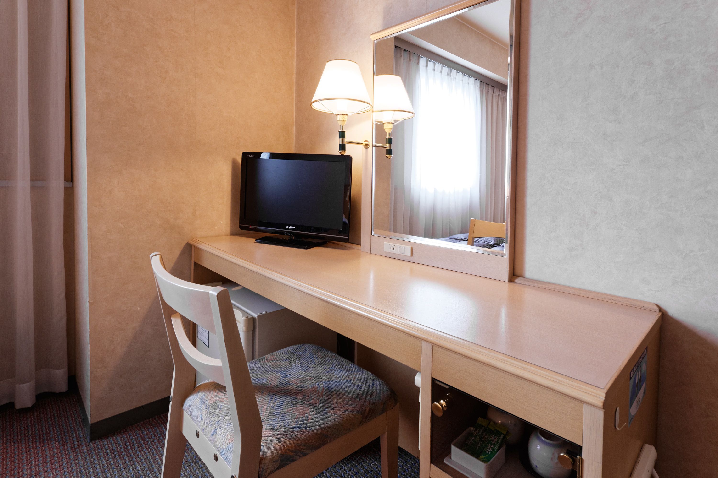 Quarto de hotel com escrivaninha de madeira, televisão, cadeira, espelho e abajur.
