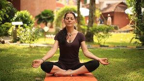 A person sitting cross-legged on an orange mat in a grassy outdoor area with trees and buildings in the background.