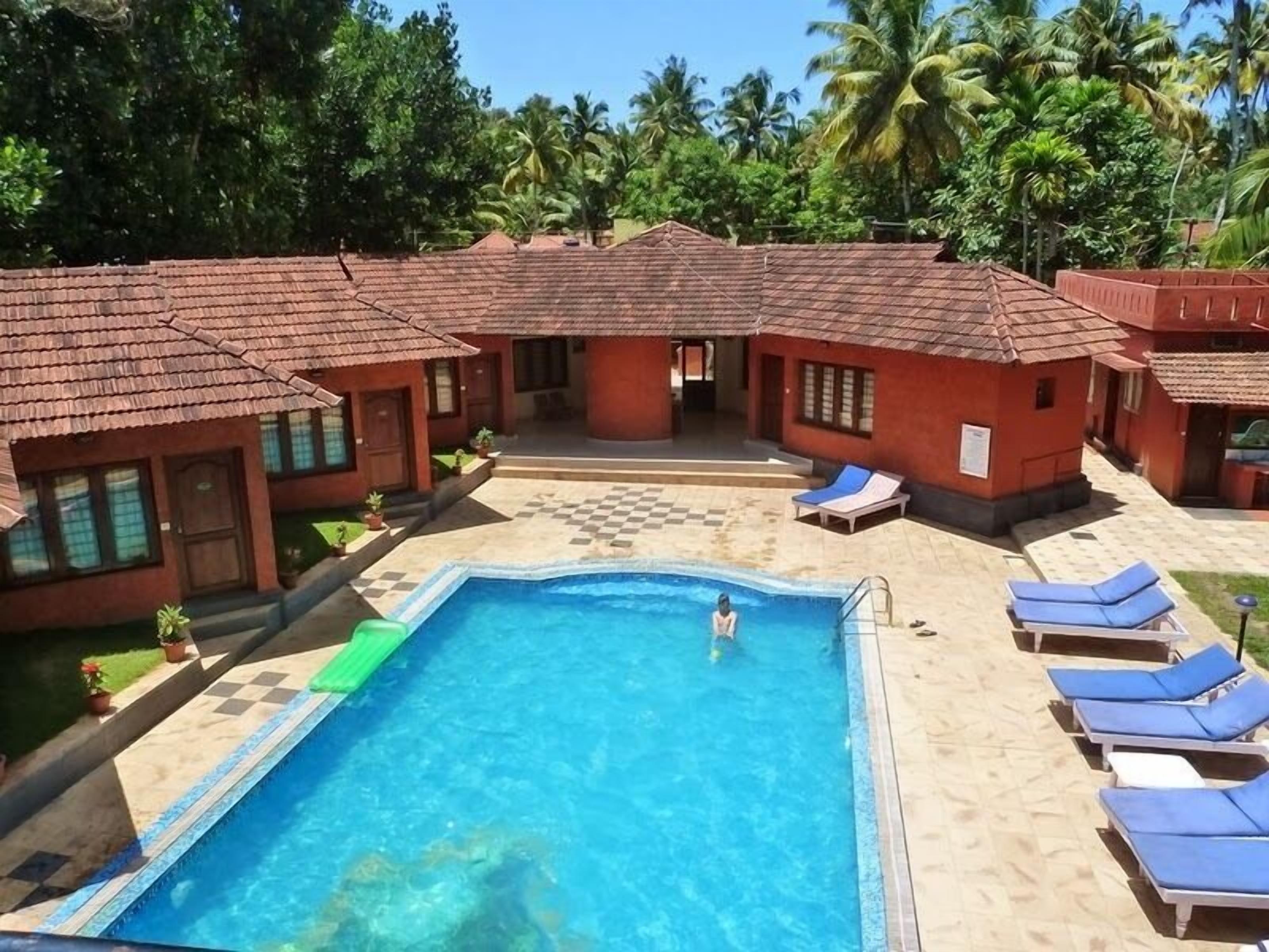 A pool area with a checkered pattern around the pool, blue lounge chairs, and red-roofed buildings.