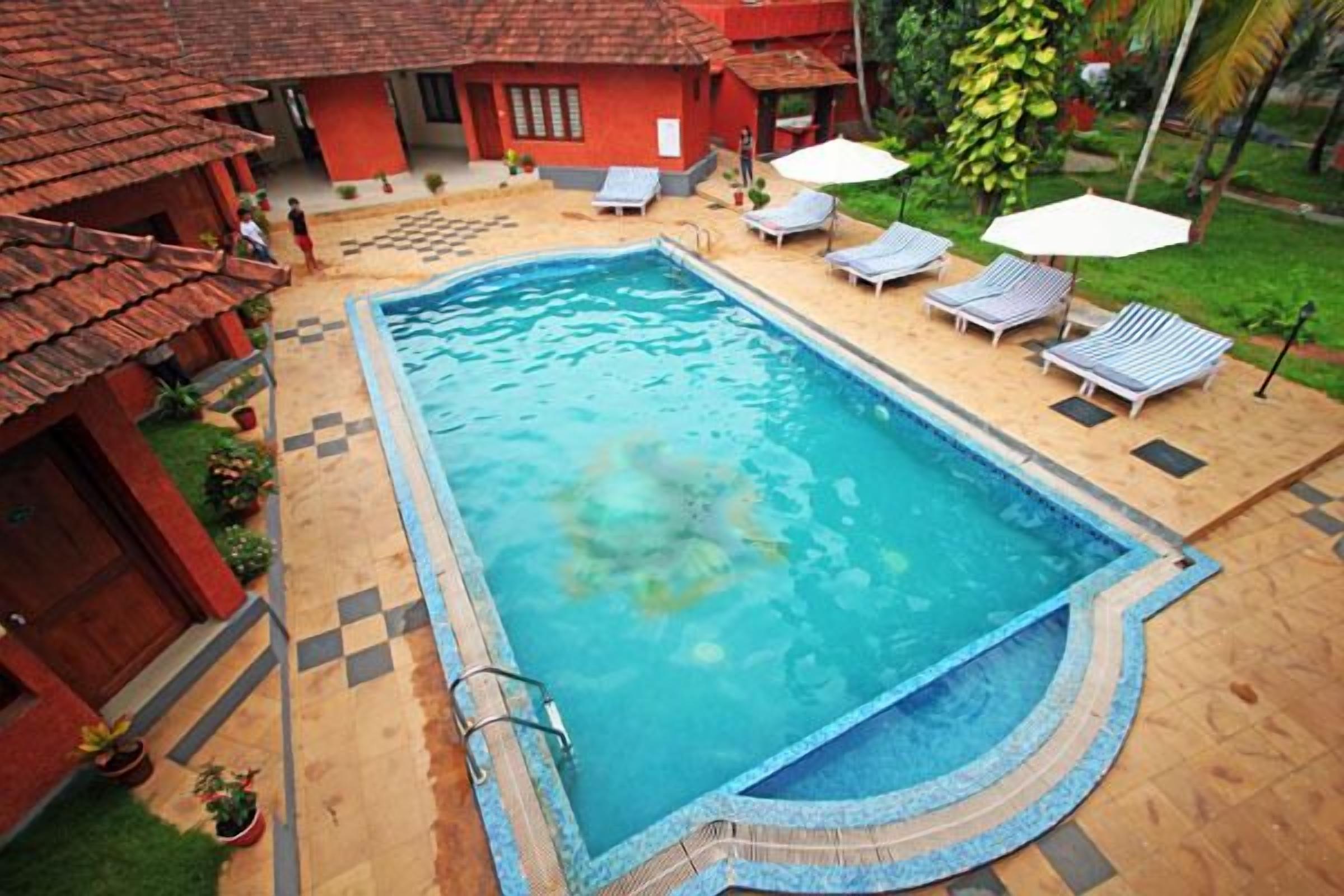 A swimming pool with lounge chairs and umbrellas, surrounded by a tiled deck and a building with a red roof.