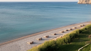Een strand met rijen ligbedden langs de kust, een rotsachtige klif op de achtergrond en een kalme zee.