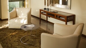 Una sala moderna con dos sillones blancos de cuero, una mesa de centro de vidrio con revistas, una mesita auxiliar de madera y un espejo grande en la pared.