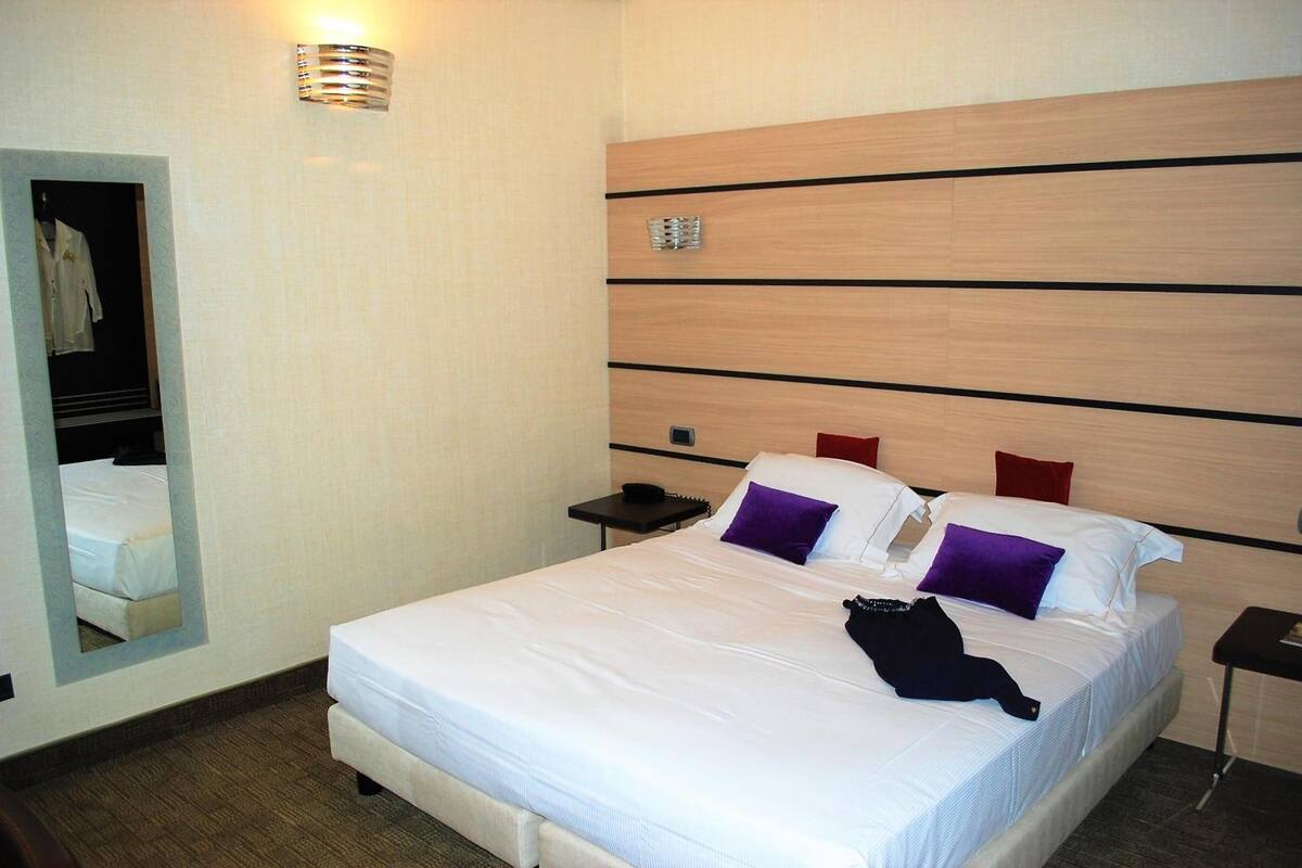 A hotel room with a large bed, a bedside table, a mirror, and a wall-mounted light fixture.