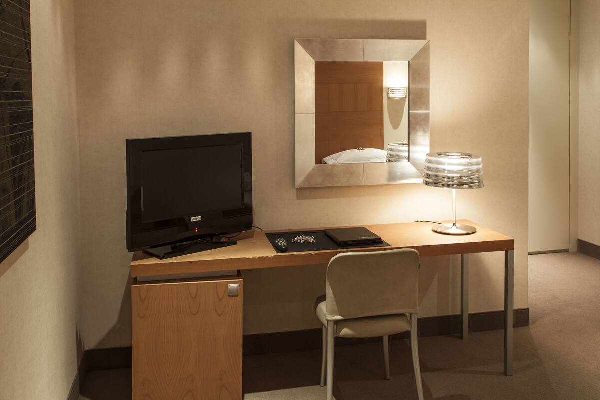 A compact hotel room with a wooden desk, a television, a chair, a small cabinet, and a mirror on the wall.