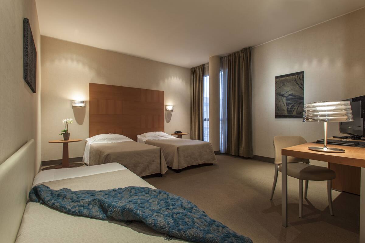 A hotel room with two beds, a desk, a chair, a lamp, and a small table with a plant.