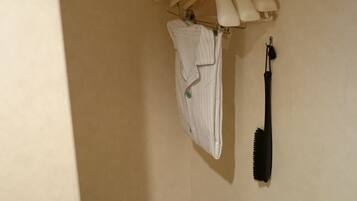 A small, dimly lit room with a single bed, a hanging clothes rack with a few garments, and a black hairbrush on the wall.