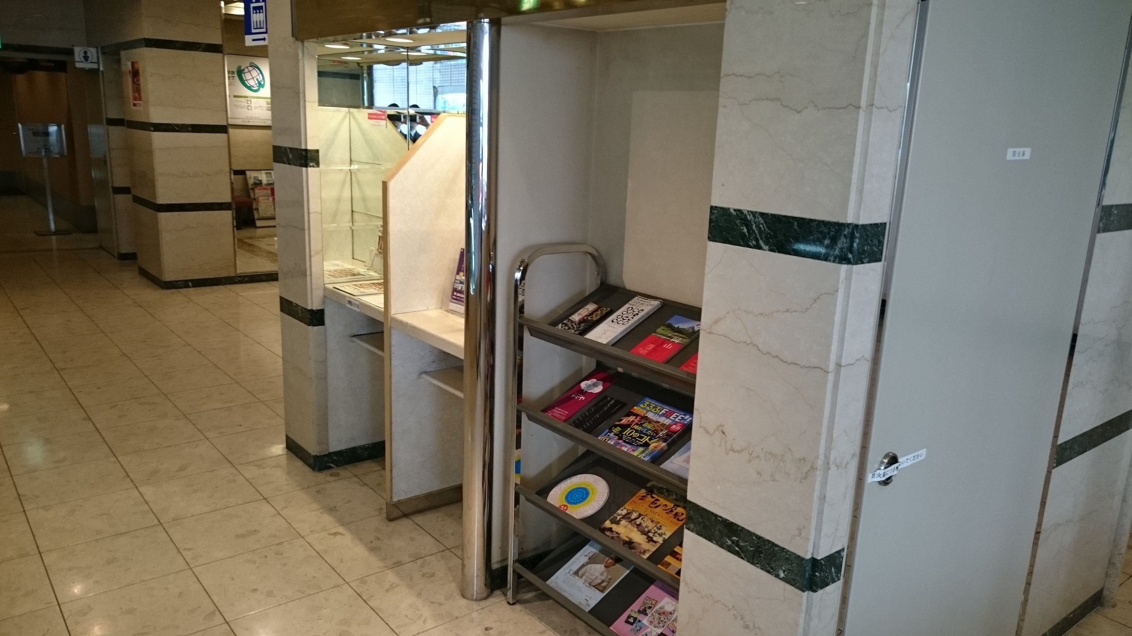 Des toilettes publiques avec un mur en marbre, une étagère en métal où sont posés des magazines et un sol carrelé.