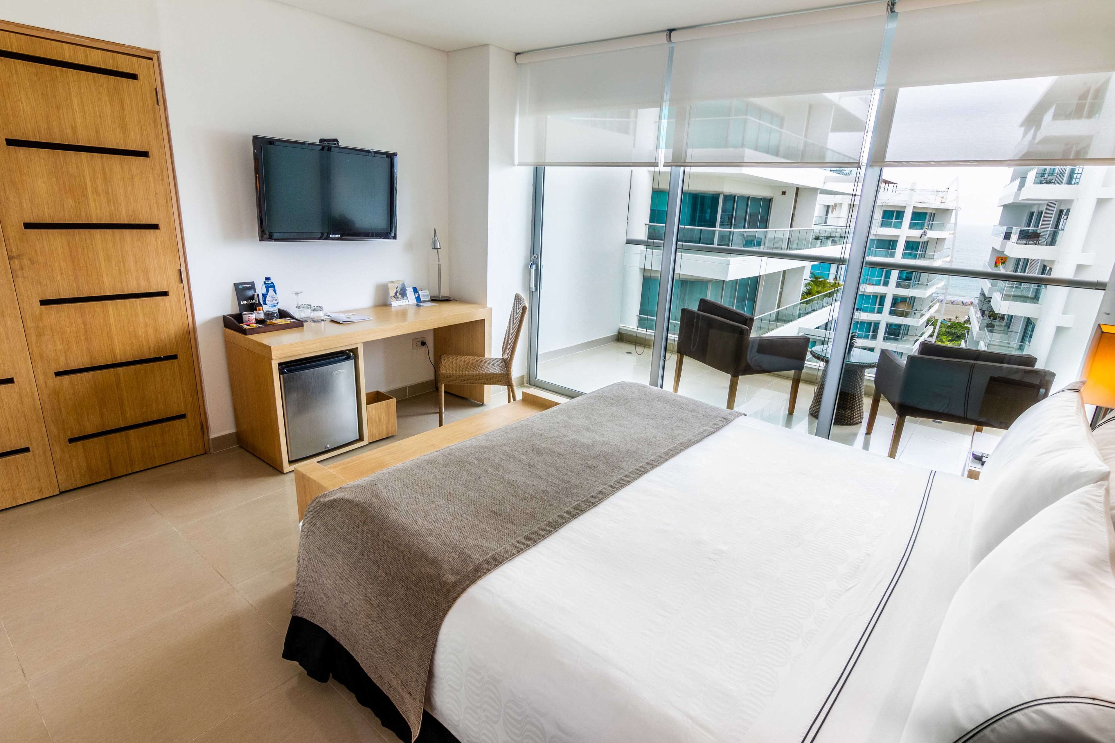 A modern hotel room with a large bed, a desk with a computer, a TV, and a balcony with a view of the city.