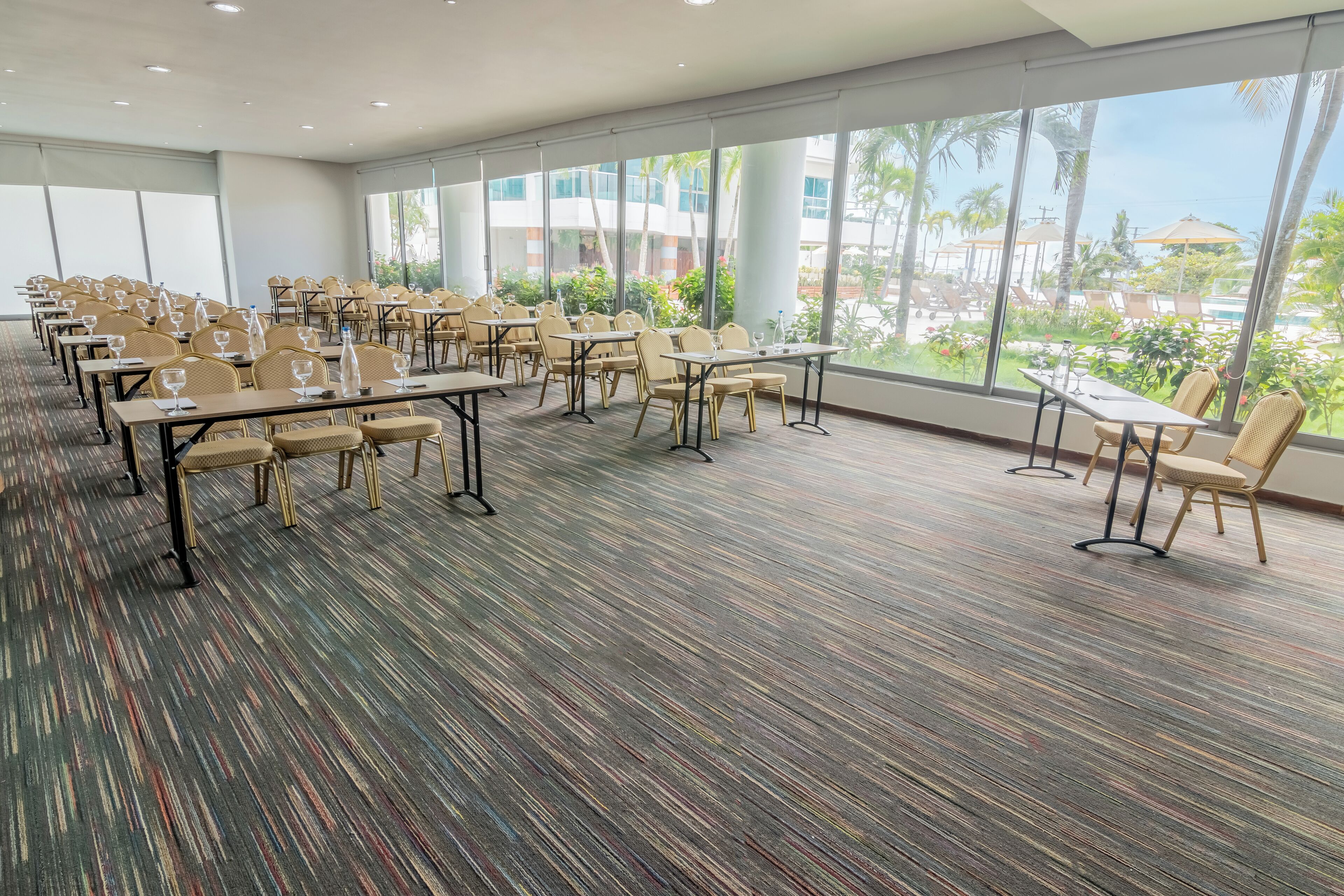 A conference room with tables and chairs, large windows, and a view of a pool and palm trees.