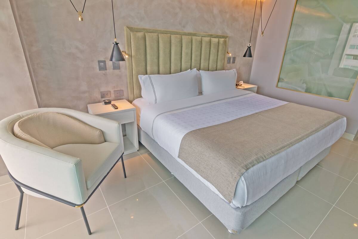 A modern bedroom with a large bed, a bedside table, a chair, and a wall-mounted artwork.