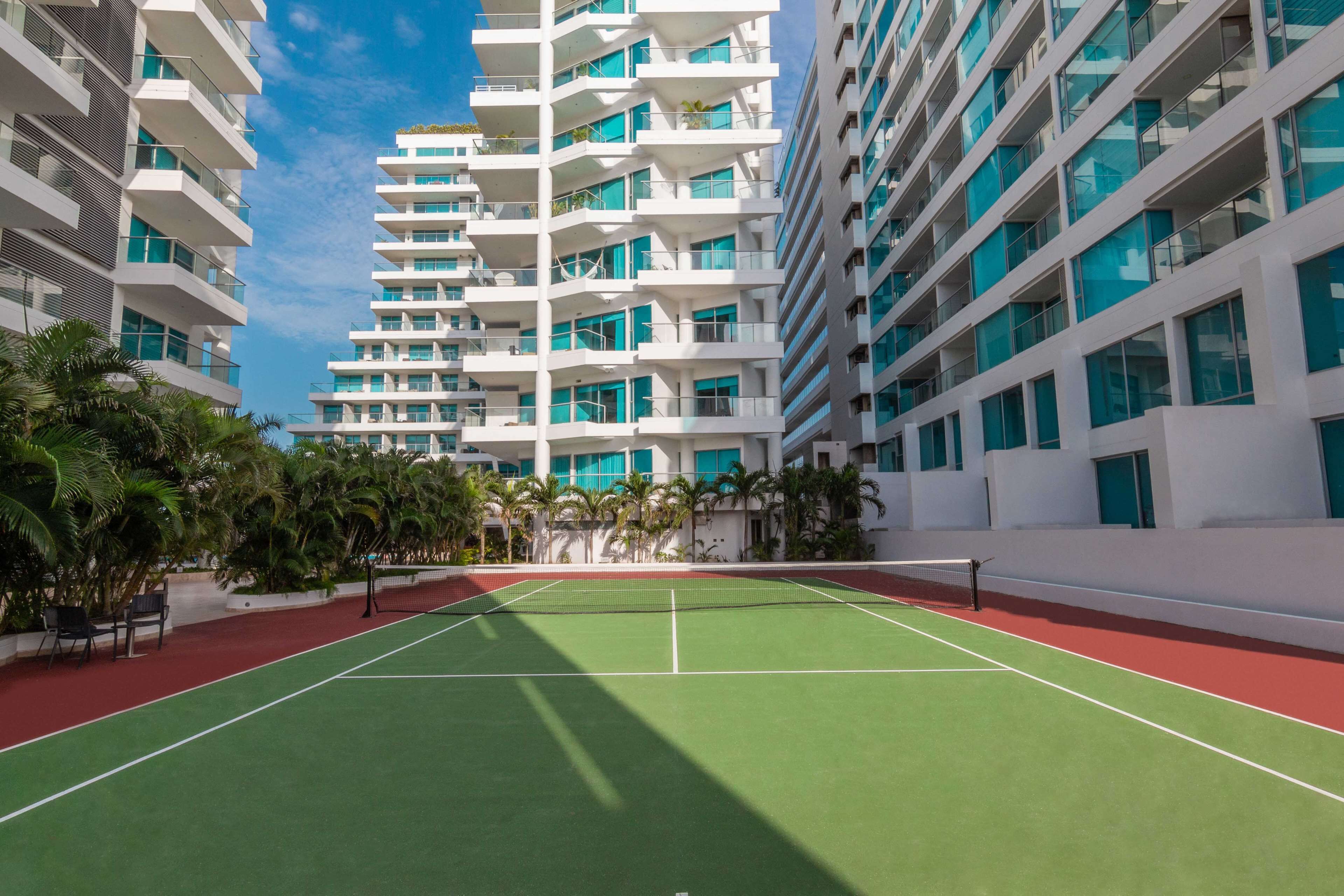A tennis court surrounded by modern buildings with balconies and large windows.