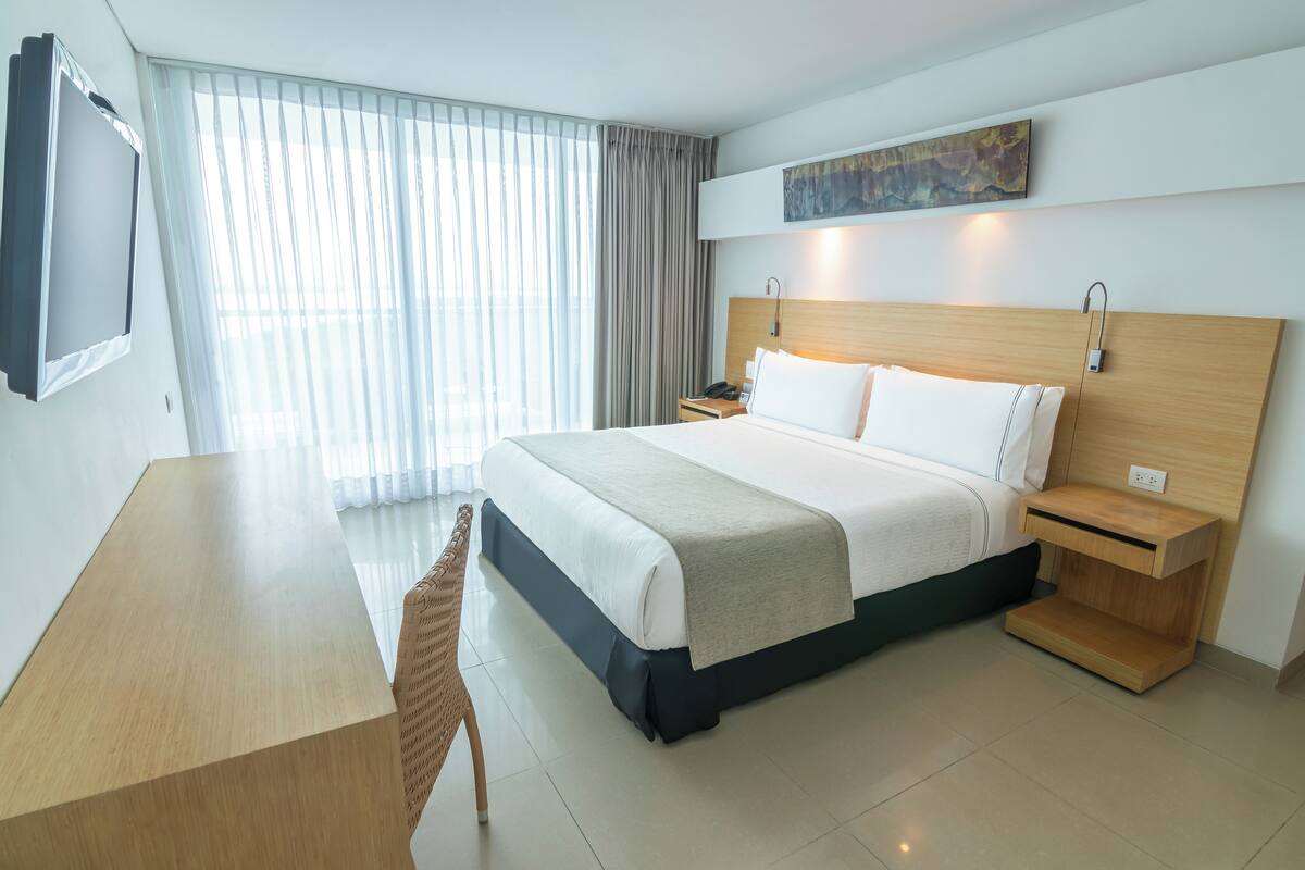 A hotel room with a bed, bedside table, lamp, and a TV mounted on the wall.