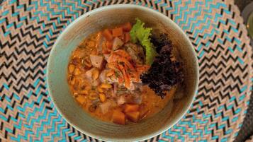Un tazón de sopa con vegetales y carne, sobre un tapete tejido.