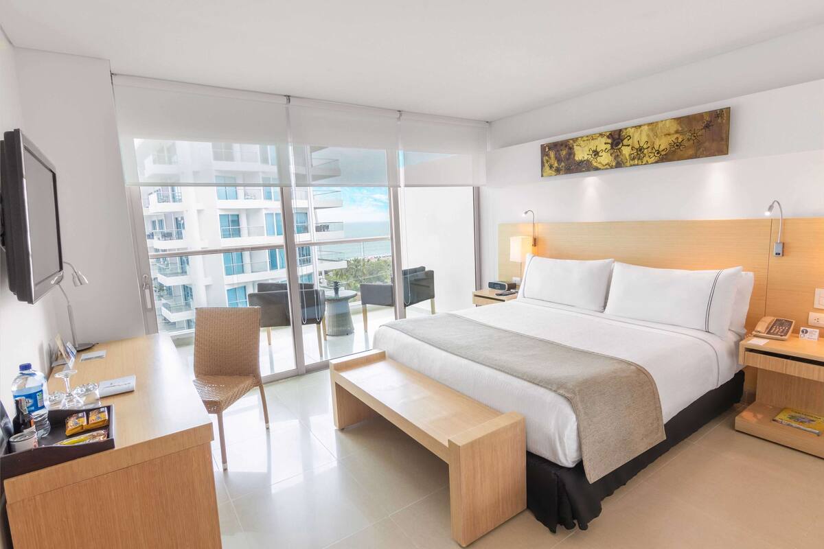 A modern hotel room with a large bed, a desk, a bench, and a view of the city through large windows.