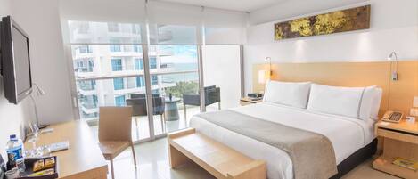 A modern hotel room with a large bed, a desk, a bench, and a view of the city through large windows.