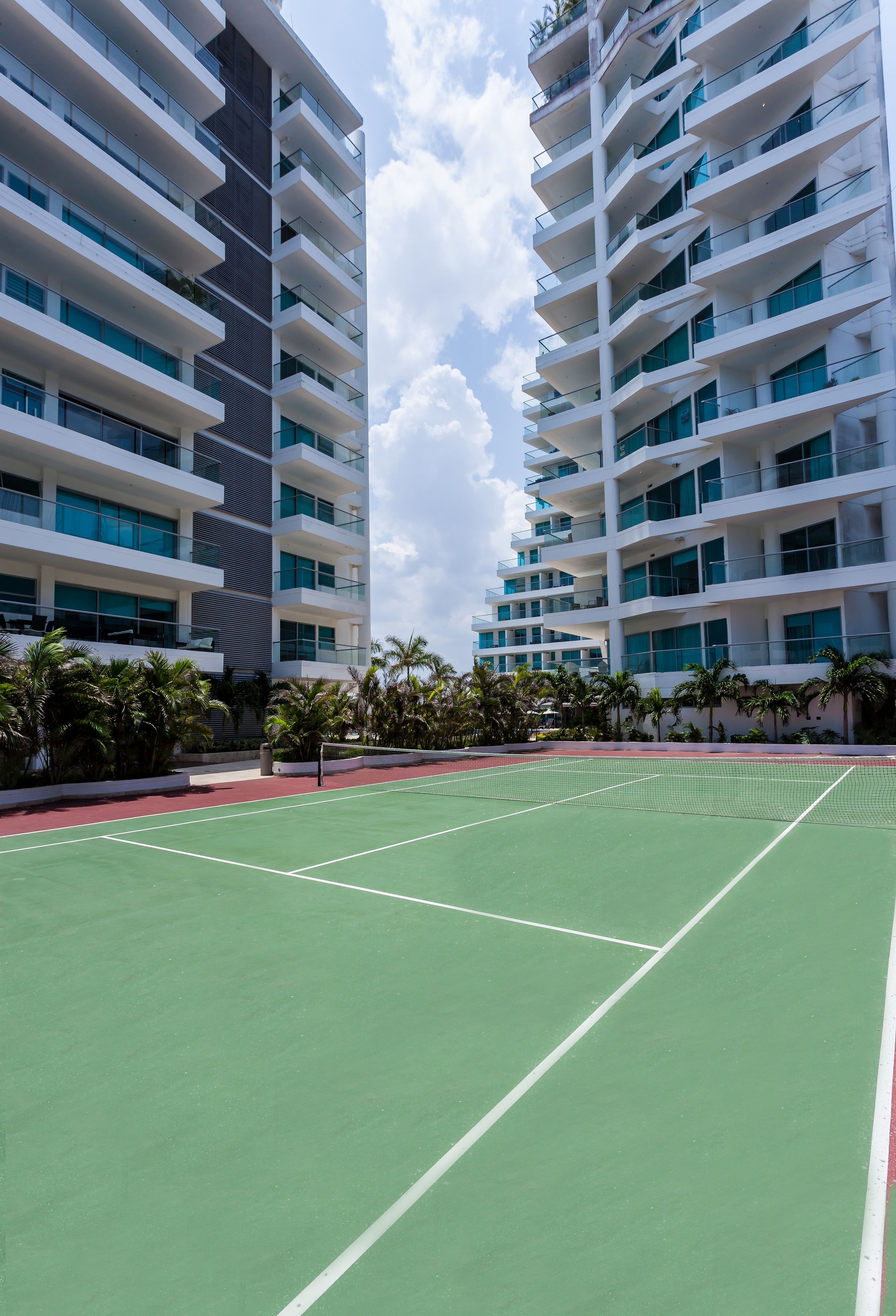 A tennis court surrounded by high-rise buildings with balconies and palm trees.