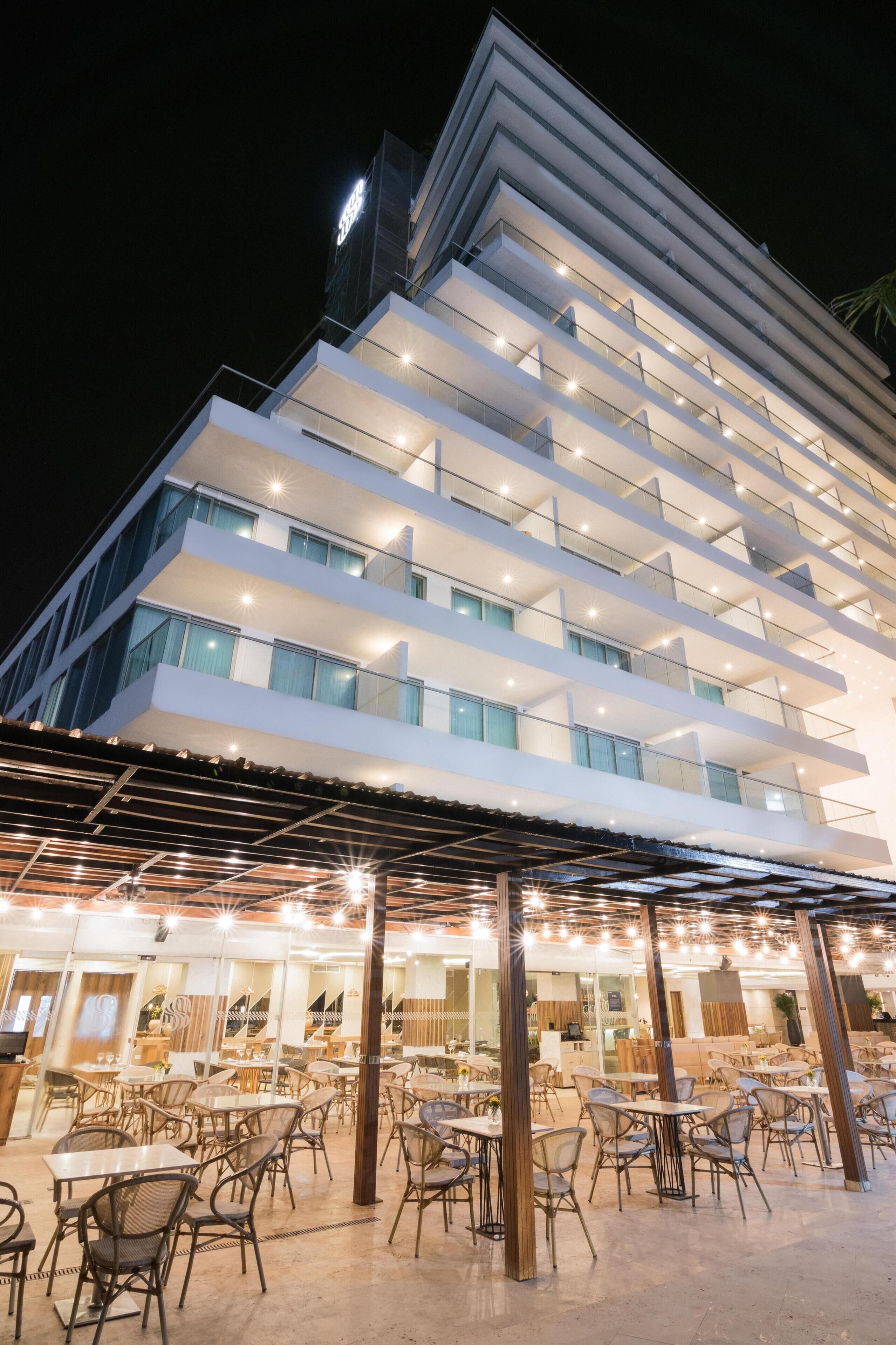 Un hôtel à plusieurs étages, doté d’un design moderne, avec des balcons et un espace de restauration extérieur couvert, comprenant des tables et des chaises.