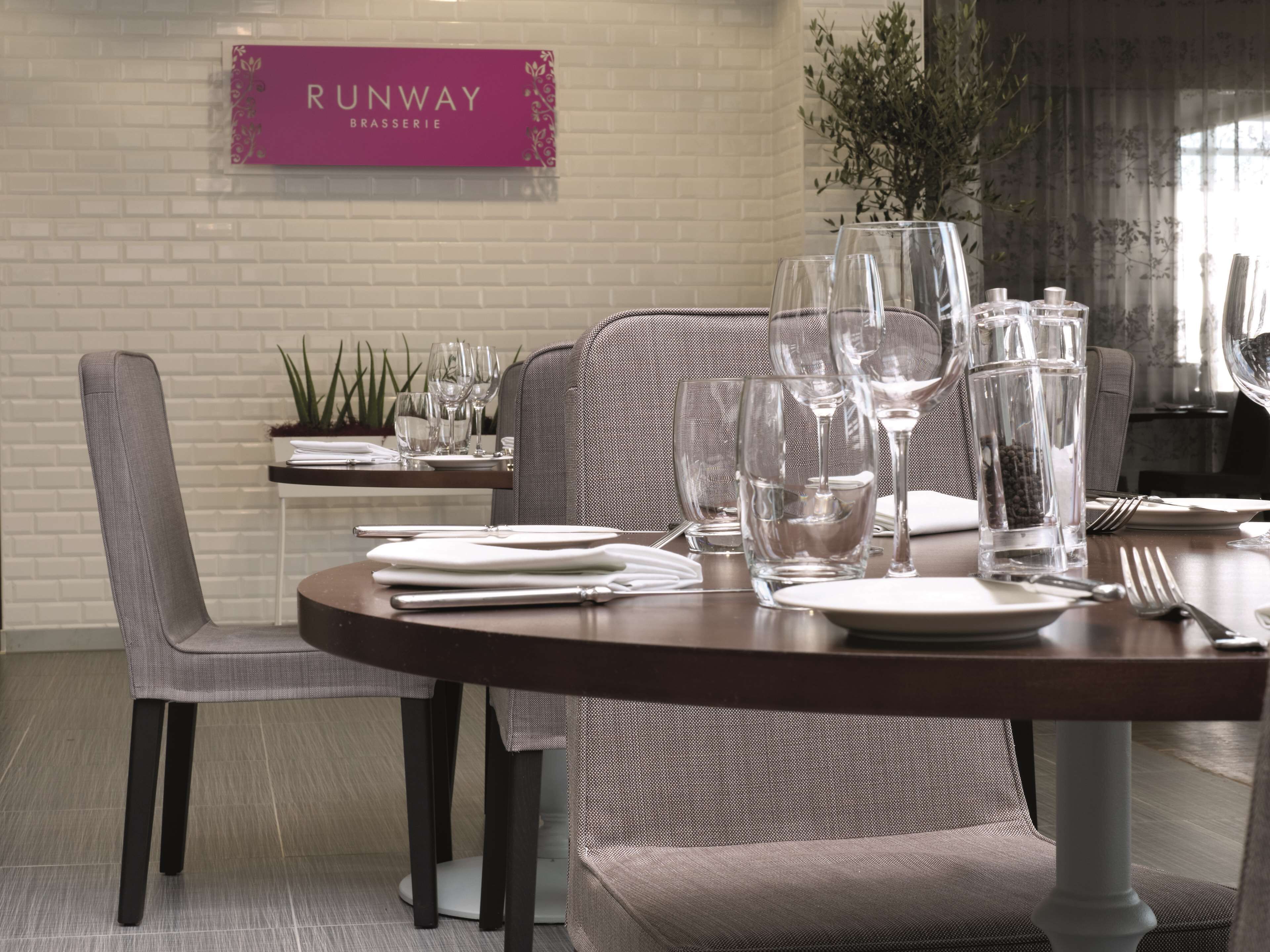 A dining area with a round table set for two, complete with plates, cutlery, and glasses. A sign above the table reads "RUNWAY BRASSERIE."