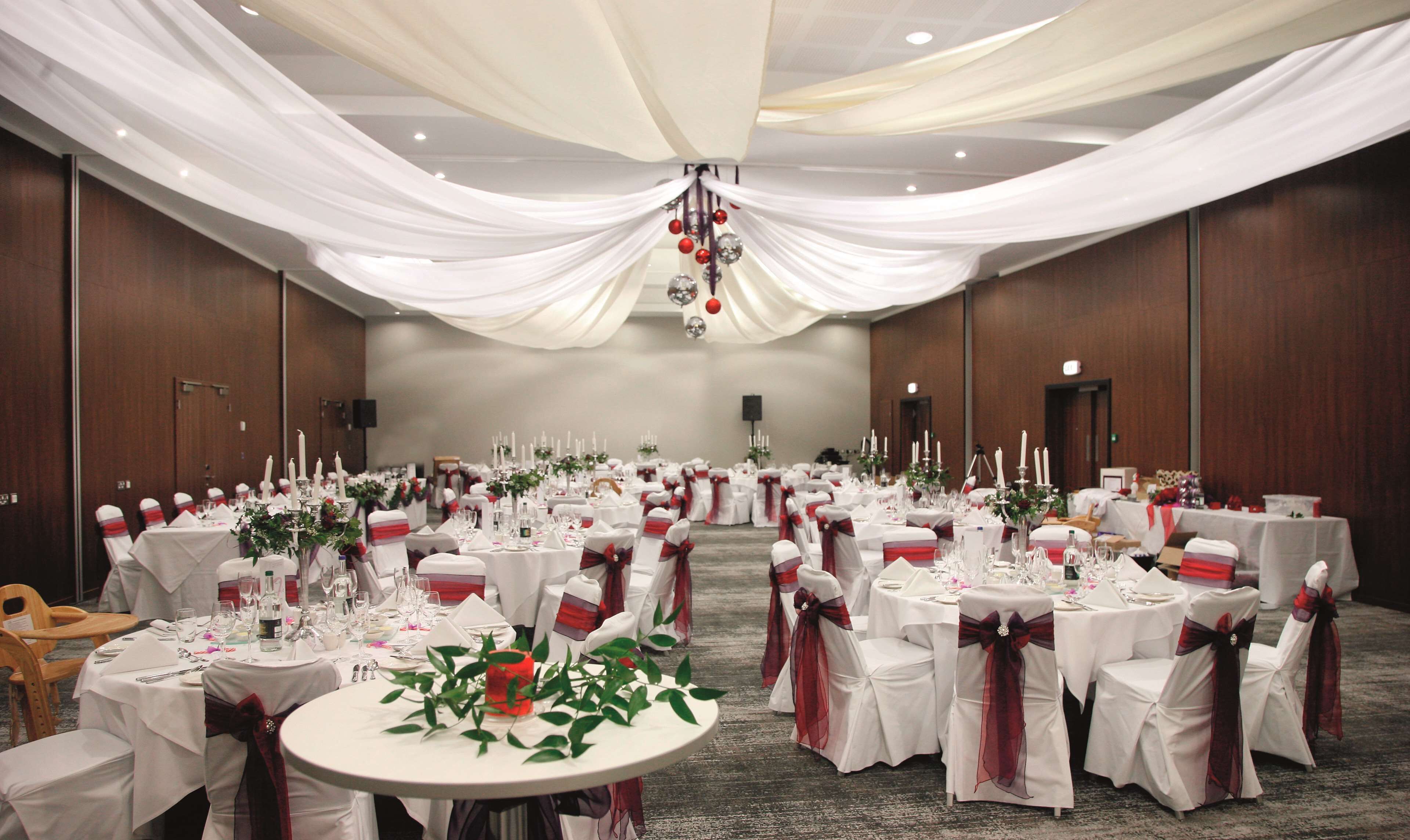 Un salón de banquetes con mesas redondas dispuestas para un evento formal, con manteles blancos y cintas rojas en las sillas.