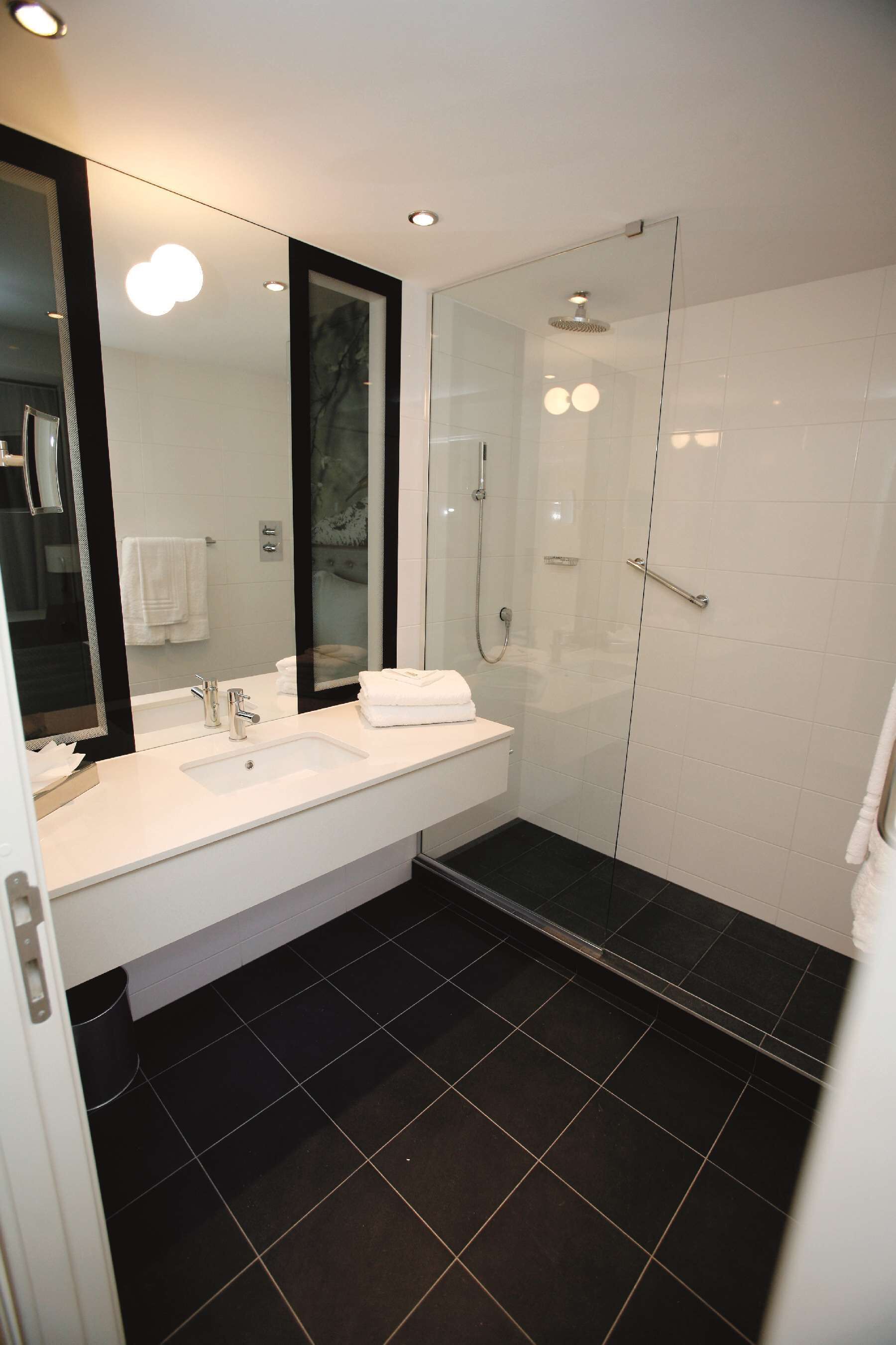 A modern bathroom with a large mirror, two sinks, a glass-enclosed shower, and black tiled flooring.