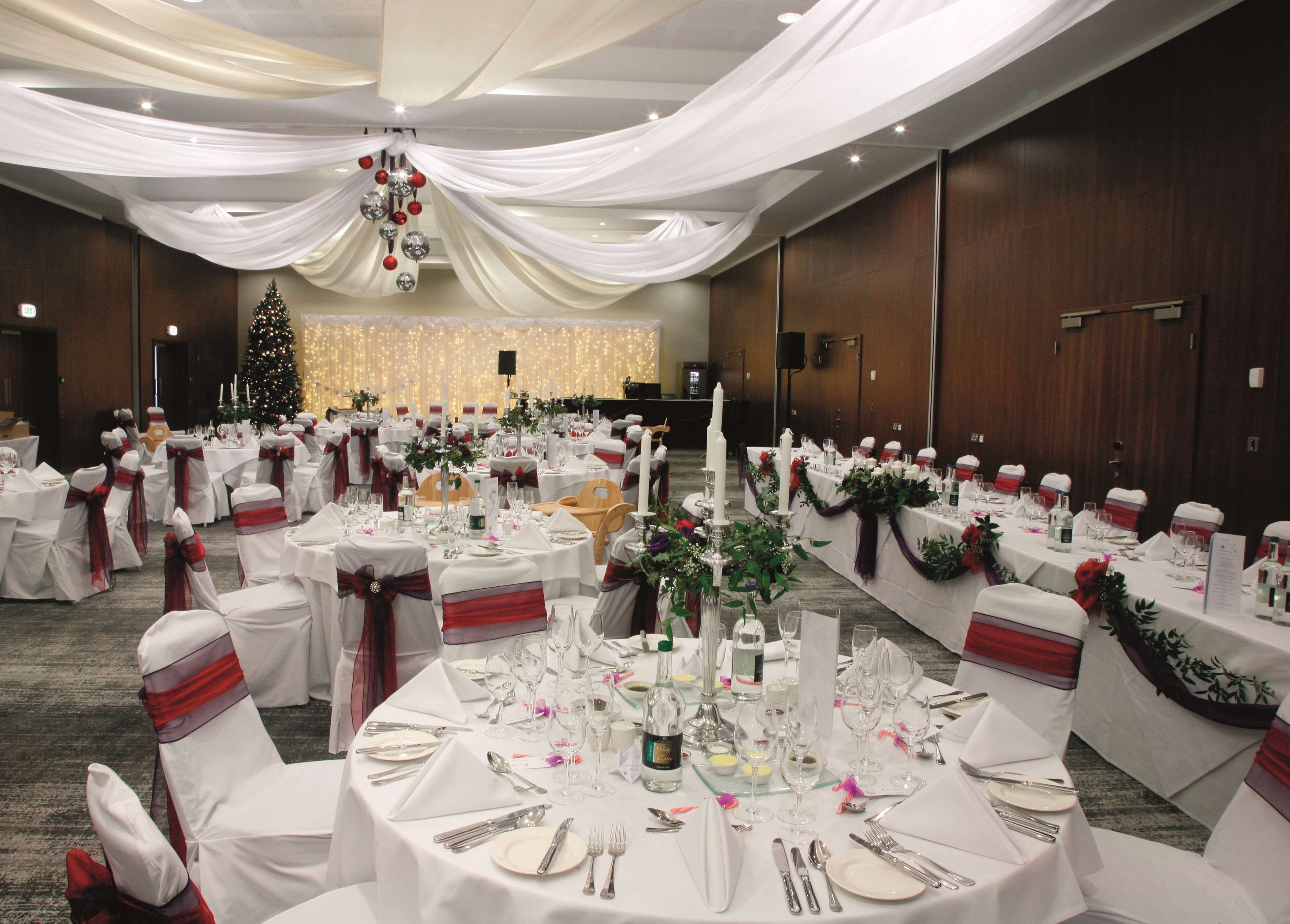 A banquet hall with round tables set for a formal event, featuring white tablecloths, red sashes, and candle centerpieces.