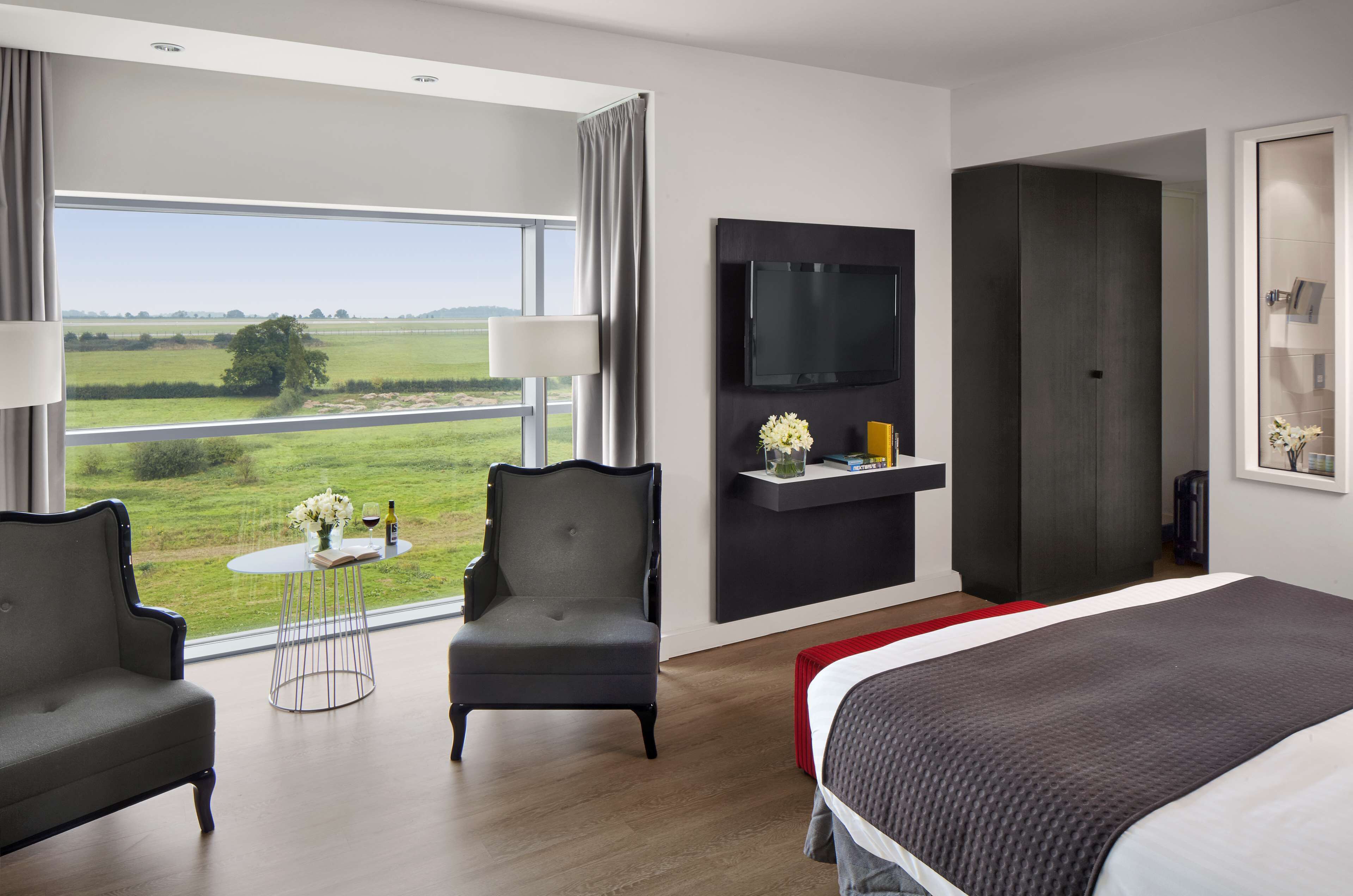 A modern hotel room with a large bed, two armchairs, a small round table with a vase and wine bottle, a flat-screen TV mounted on the wall, and a large window offering a view of the countryside.