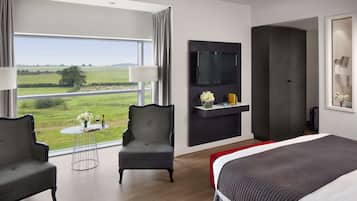 A modern hotel room with a large bed, two armchairs, a small round table with a vase and wine bottle, a flat-screen TV mounted on the wall, and a large window offering a view of the countryside.