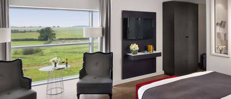 A modern hotel room with a large bed, two armchairs, a small round table with a vase and wine bottle, a flat-screen TV mounted on the wall, and a large window offering a view of the countryside.
