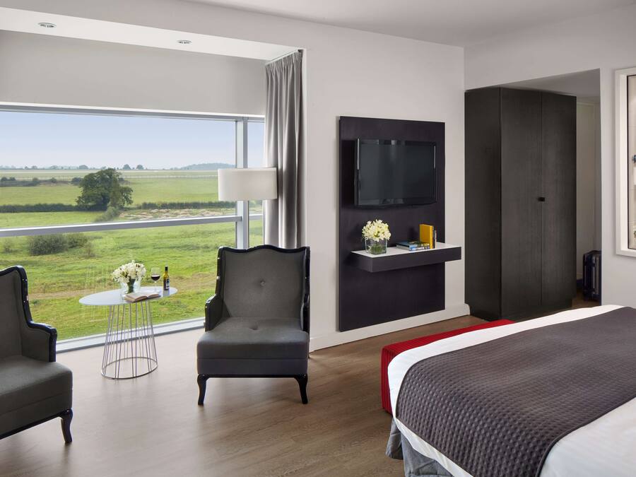 A modern hotel room with a large bed, two armchairs, a small round table with a vase and wine bottle, a flat-screen TV mounted on the wall, and a large window offering a view of the countryside.