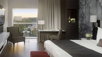 Habitación de hotel moderna con una cama grande, un escritorio, dos sillas, un televisor y vistas a un paisaje urbano.