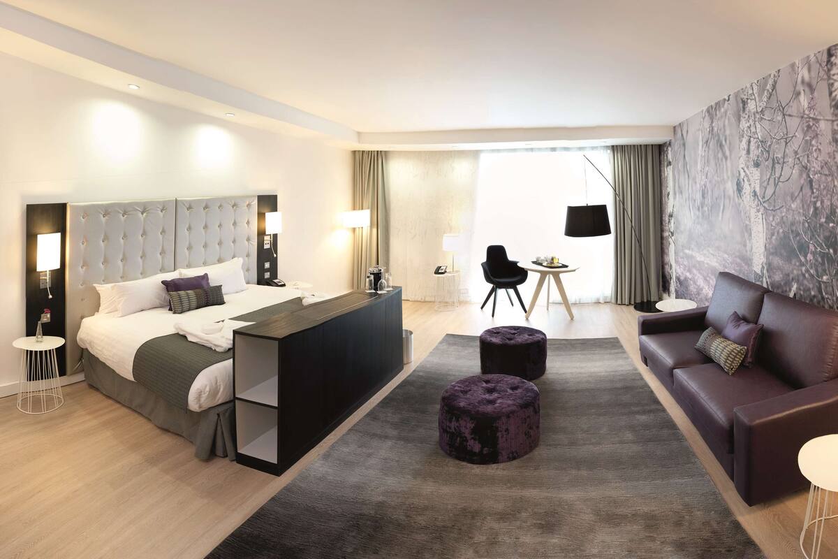 A modern hotel room with a large bed, a desk, a sofa, and a dining area.