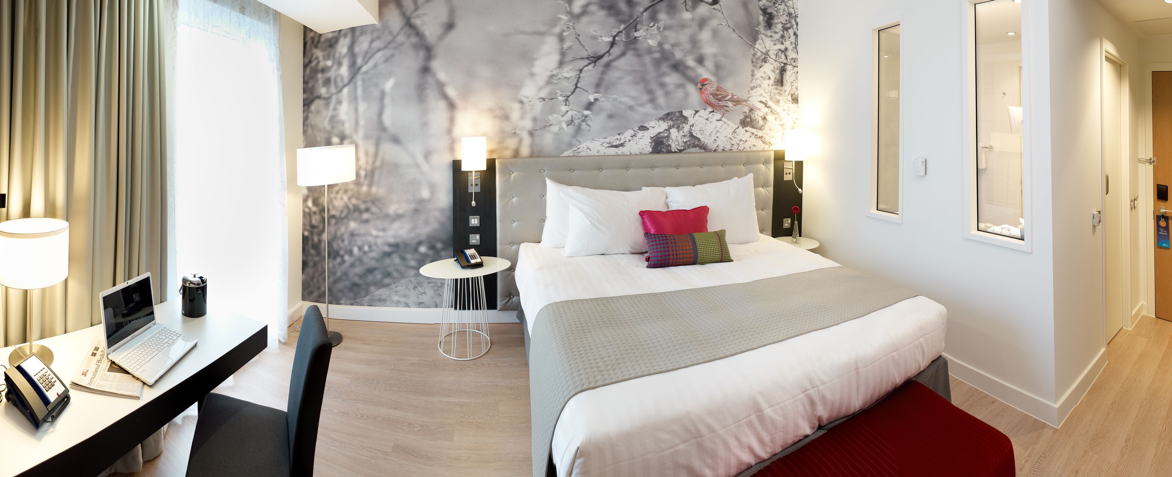 A modern hotel room with a bed, a desk with a laptop, a chair, a small round table, and a wall with a tree mural.